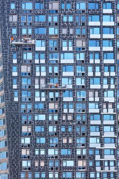 Several teams of builders, being on construction cradles, insulate the facade of a residential building under construction, the background and texture of glass windows, a vertical image.