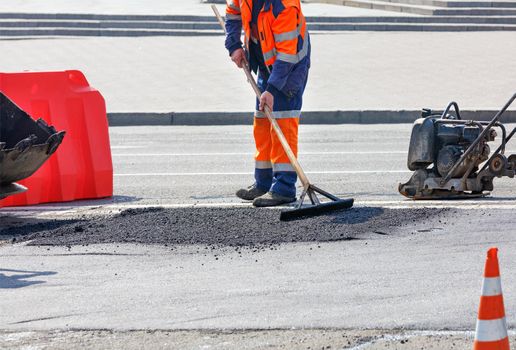 A road worker in a bright orange overalls on a fenced-off work section of the road patches a pothole with hot asphalt and ramps it with a petrol vibratory plate.