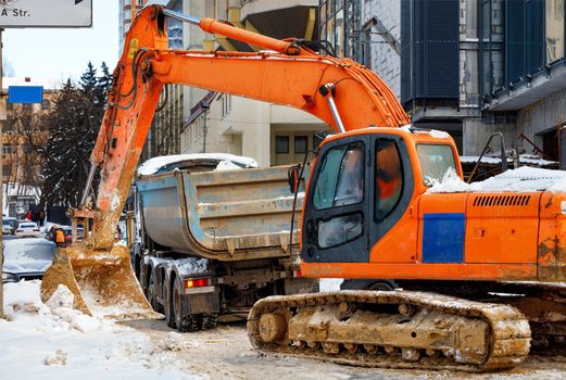 A large orange crawler excavator loads snow from a city road onto a truck bed with its bucket in front of a city street.