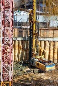 A large crawler excavator installs concrete columns with a drilling rig to strengthen the foundation of a future high-rise, viewing angle from a height, sunny day, vertical image.