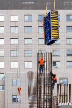 Workers in bright orange vests and safety helmets are using a crane at a height to mount the formwork on a metal frame made of steel reinforcement against the background of the window facade of the new house. Vertical image.