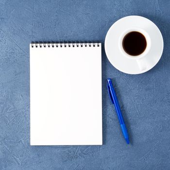 The open notepad with clean white page, pen and coffee cup on aged dark blue stone table, top view