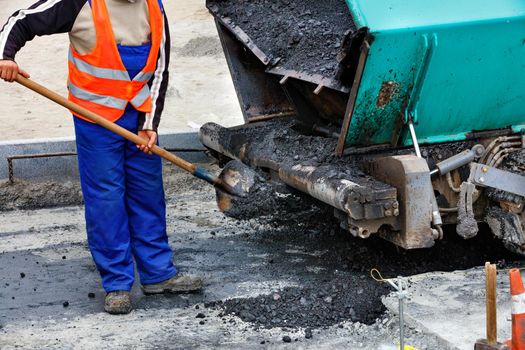 A worker in a blue overalls and an orange jacket cleans an asphalt paver with a shovel. Selective focus.