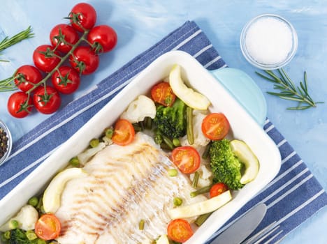 Fish cod baked in blue oven with vegetables - broccoli, tomatoes. Healthy and diet food. Blue stone background, top view.