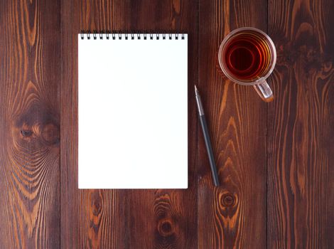 The open notepad with spiral , pencil, cup of tea on a dark brown wooden table, top view