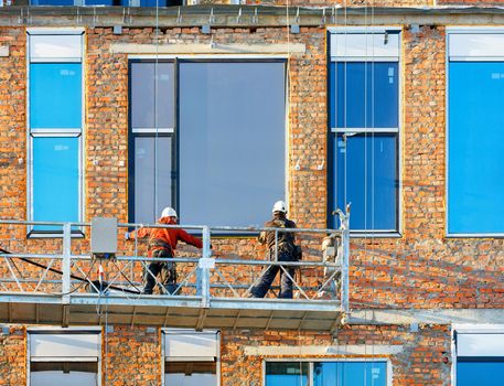 Builders install large windows on the facade of a house under construction using a cradle and an aluminum profile, which is a concept for saving heat in the house.