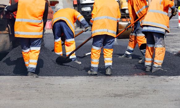 A team of road workers in orange reflective uniforms repair a section of road with fresh asphalt. Clear noon, copy space.