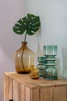 Transparent yellow and green vases for flowers and big bottle with monstera leaf on wooden cupboard. Decoration of interior