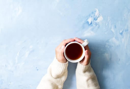 Woman holding cup of hot tea on a blue table, hands in warm sweater with mug, winter morning concept, top view