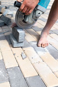 A worker with a grinder with a diamond cutting wheel trims paving slabs, standing barefoot and holding a stone cobblestone with one foot. Copy space, vertical shot.