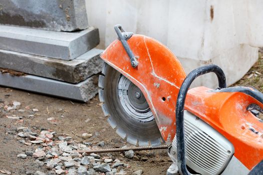 A worn, bright orange gasoline cutter with a diamond cut-off wheel set against a background of concrete parapets and rubble in blur.