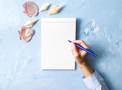 woman writes in notebook on a stone blue table, Mock up with frame of seashell, top view, planning holiday by sea