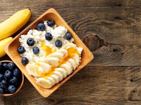 Brown wooden Bowl of homemade curd with banana, jam, blueberries on a dark brown wooden background, top view, copy space