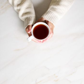 Woman holding cup of hot tea on stone beige marble table, closeup photo of hands in warm sweater with mug, winter morning concept, top view