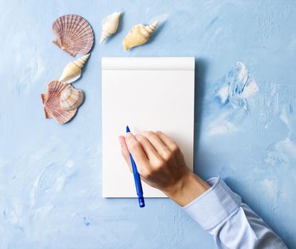 woman writes in notebook on a stone blue table, Mock up with frame of seashell, top view, planning holiday by sea