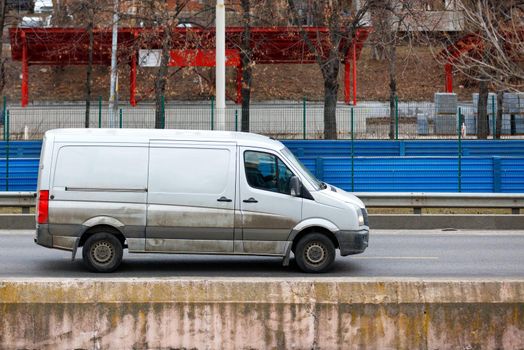 A white cargo van with muddy lower sides moves along a city street highway on a spring afternoon.