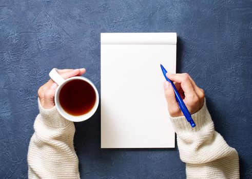 woman writes in notebook on a dark blue table, hand in shirt holding a pencil, cup of tea, sketchbook drawing, top view