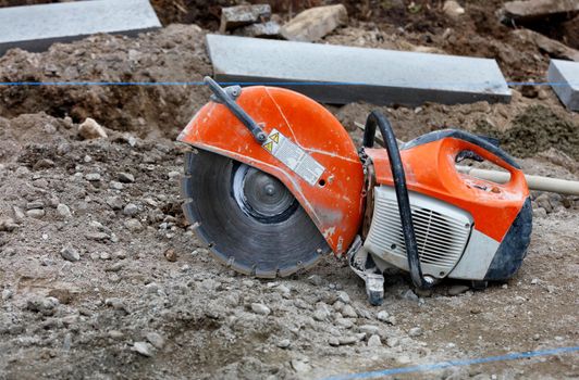 A worn, bright orange petrol saw with a diamond cut-off wheel set against a blurred background of rubble, concrete parapets and taut blue thread.