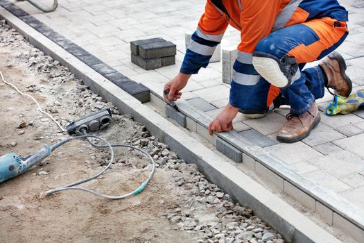 A bricklayer in a blue-orange overalls, half-squatted, measures out a part of the paving slabs with a black marker and an aluminum level for subsequent even laying. Close-up, selective focus, copy space.