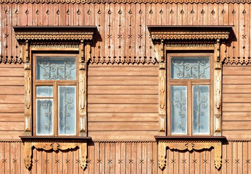 The old wooden textured facade of the house with carved platbands, curly wall elements and metal bars in the windows, illuminated by warm sunlight.