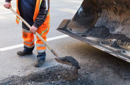 A road service worker shovels old asphalt into the metal bucket of an excavator while repairing a section of the road.