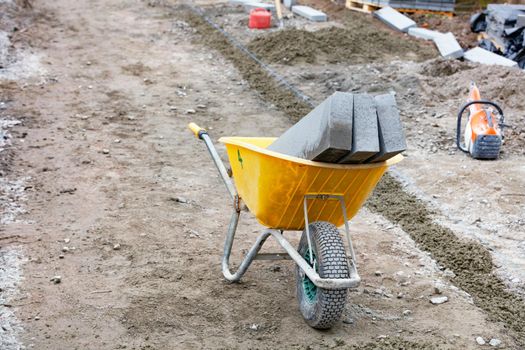 Construction yellow wheelbarrow with loaded curb concrete blocks at a marked sidewalk repair construction site on a spring afternoon. Copy space.