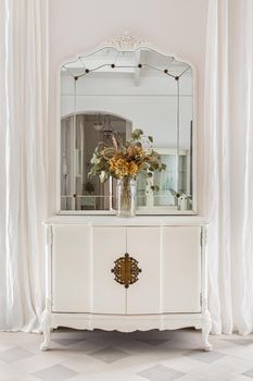 Old restored furniture in classic bright interior with white wall and curtains. Vintage mirror and flower bouquet on wooden cupboard