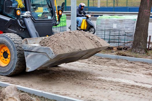 A compact grader on a construction site carries a sand bucket to level the sandy base on the sidewalk under construction. Subtle motion blur, copy space.