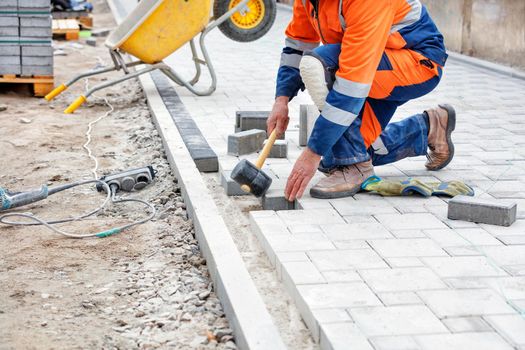 A bricklayer in a blue and orange overalls, half-squatting, taps the paving slabs with a black rubber mallet for subsequent smooth installation. Close-up, selective focus, copy space.