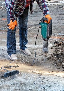 A construction worker in a plaid work shirt and blue jeans loosens dense soil with an electric jackhammer at a construction site, copy space, vertical image, close-up.