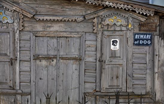 Old dilapidated wooden facade of the house with carved platbands, curly wall elements and metal bars, with a sign Beware of dog.