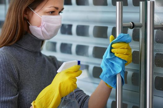 Female cleaning lady, in protective mask and gloves, wipes door handles with antiseptic in restaurant or public establishment. Protection against covid-19, safety concept