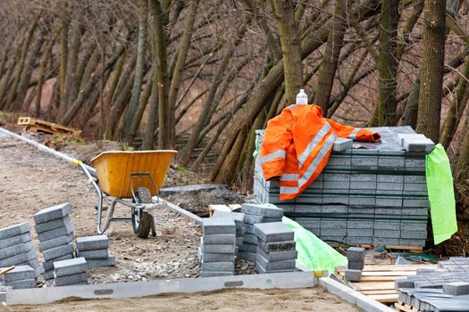 Construction site with a stack of paving slabs and an orange builder jacket near a yellow wheelbarrow against the backdrop of park trunks of growing trees on a spring day.