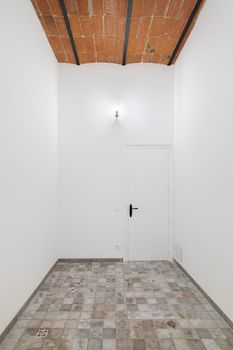 Empty white narrow room with closed door, spotlight, tiles floor and vintage brick ceilings.