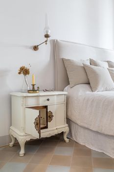 Interior of cozy house in retro style. Vertical photo of classic bedroom with candle, glasses and flowers on wooden bedside table near comfortable bed