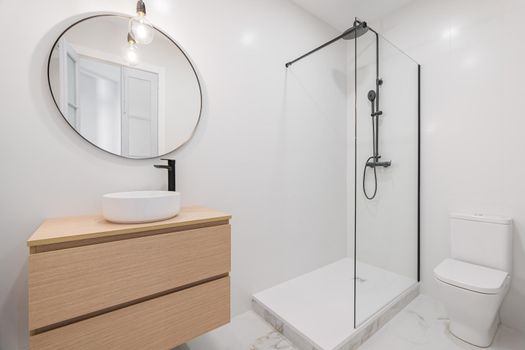 Interior of white bathroom in modern design, with shower zone, toilet, marble floor, wooden furniture and round mirror.