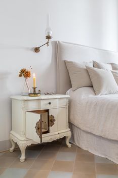 Interior of cozy house in retro style. Vertical photo of classic bedroom with candle, glasses and flowers on wooden bedside table near comfortable bed