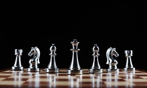 Shiny steel chess figures standing on wooden chessboard. Intellectual duel and tactical battle symbol. Strategy planning and corporate leadership concept. Silver metal chess pieces on black background