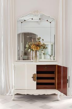 Vintage mirror and flower bouquet on wooden cupboard. Old restored furniture with opened door and drawers in classic bright interior with white wall and curtains.