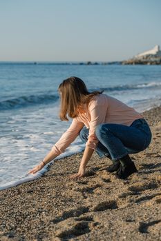 Young woman in pink shirt and blue jeans touching the wave with hand at the sea beach