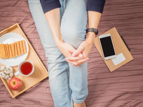 Flatlay of woman in jeans sitting on her bed hugging her knees, with smartphone, notepad, breakfast with tea, top view.