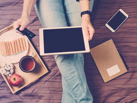 A woman sitting on a bed, drinking tea and looking at tablet, pays for shopping, online shopping. Tablet with dark screen