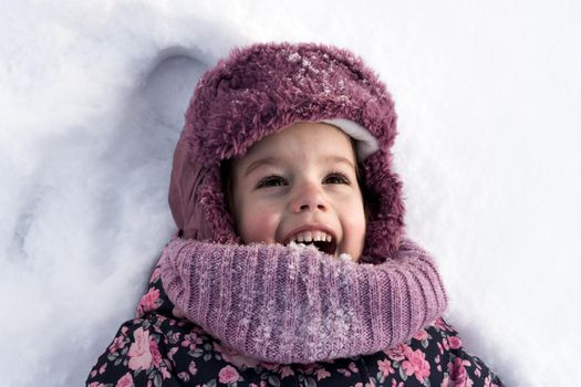 Winter, family, childhood concepts - close-up portrait authentic little preschool girl in pink clothes smile laugh shout with open mouth laying on snow in frosty weather day outdoors. Funny kid face.