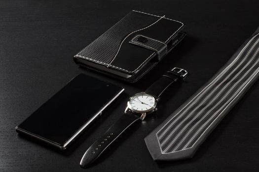 Watch with a leather strap, notebook in leather cover, sell phone, silk tie on a black background