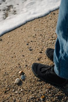 Legs of a person in black shoes and blue jeans standing at beach with sea wave at the sunset.
