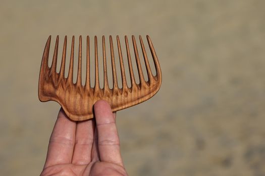 A hand holding natural wooden comb made of pear tree for scalp massage, aroma combing at sand background.