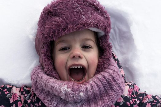 Winter, family, childhood concepts - close-up portrait authentic little preschool girl in pink clothes smile laugh shout with open mouth laying on snow in frosty weather day outdoors. Funny kid face.