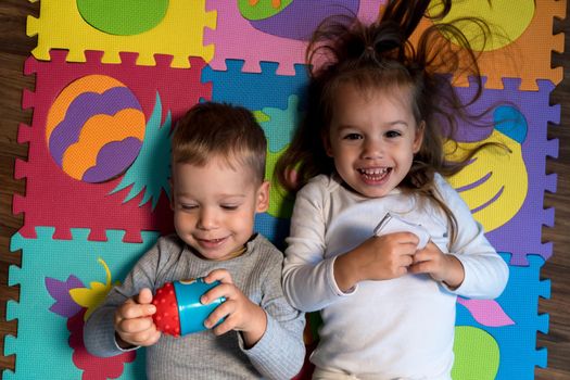 childhood, family friendship, games - close up portrait Two funny joy happy smiling little toddler peschool kids siblings twins brother with sister have fun lie playing on puzzles mat at home indoors.