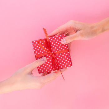 Two women holding a gift in a red gift box with a bow, passing it from one to the other, pink background, top view.