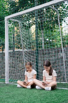 two pretty girls in identical clothes using phone on sports ground or athletic field. kids are surfing in internet. smartphone addiction. advanced, modern children in social networks.